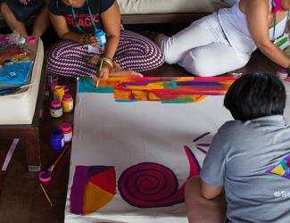 A group of people are sitting on the floor and painting together on a large canvass. There are paint containers, brushers and other craft materials to the left.
