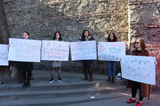 La foto presenta a cinco mujeres (Sopo está en el medio) de pie en lo alto de una escalera, frente a un muro de piedra, sosteniendo carteles en los que están escritos eslóganes georgianos.