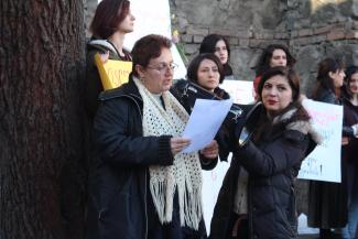 La foto muestra a ocho mujeres de pie juntas durante una protesta. Muchos sostienen pancartas mientras Sopo acerca el megáfono a la boca de una trabajadora de pelo corto y rojo, con bufanda blanca y abrigo negro, que lee un manifiesto.