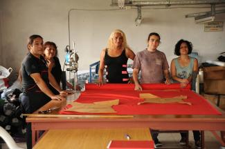 Photo of five members of the Nadia Echazu cooperative standing around a table in their workshop with a bright red piece of cut fabric spread across the table. In the middle is Brisa Escobar, the president of the Nadia Echazú cooperative