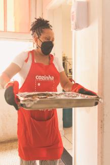 Woman in a white t-shirt, striped pants, red apron, and black facemask holding batch of food