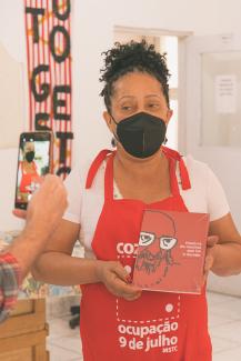 Photo of a black woman in a red apron and black facemask, holding a book