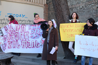Quatre personnes avec des affiches lors d'une manifestation et, au milieu, une femme avec un mégaphone qui parle. 