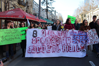 La imagen muestra una protesta en la que una multitud sostiene una pancarta en georgiano que dice: "8 de marzo para las mujeres trabajadoras".
